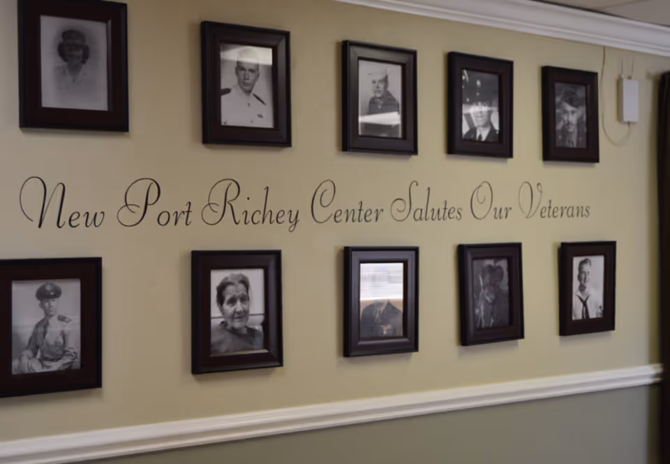 A wall display featuring framed black and white photographs of veterans, arranged in two rows. In the center of the wall, decorative text reads 'New Port Richey Center Salutes Our Veterans.' The wall is painted in a light color with a white chair rail molding below the photos.