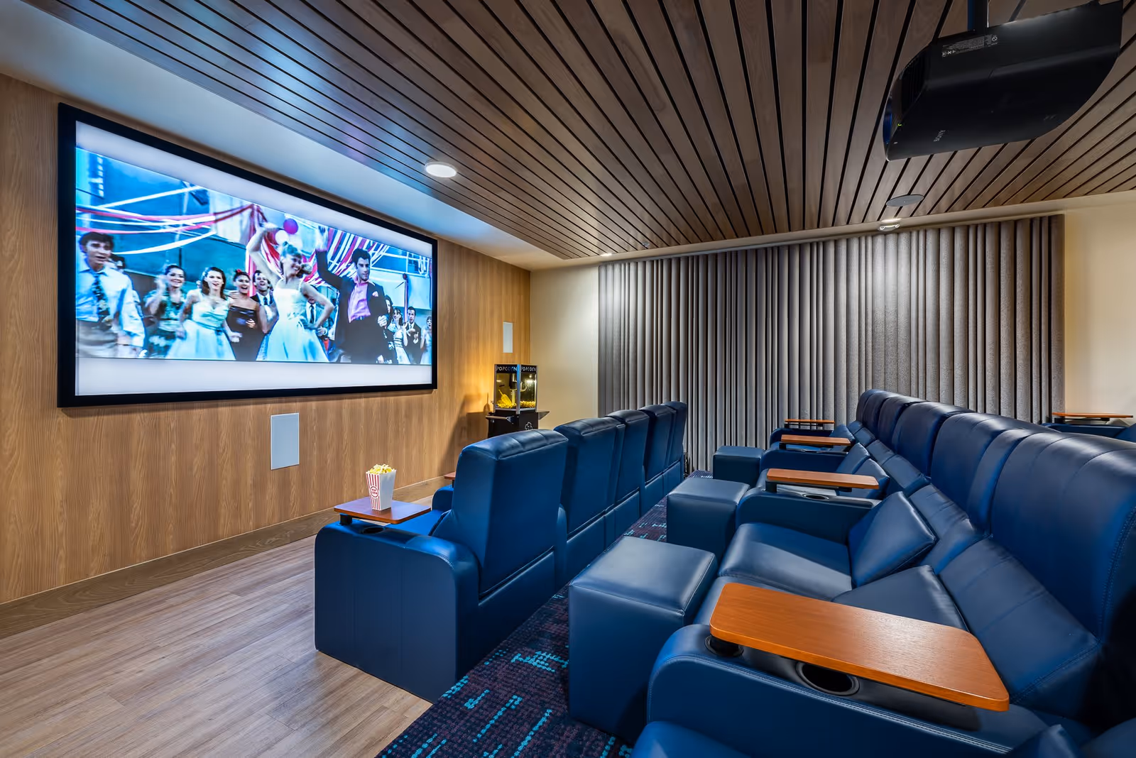 A modern home theater room with blue leather recliner seats arranged in two rows facing a large screen displaying a movie scene. The room features a wooden slatted ceiling, wood-paneled walls, a popcorn machine in the corner, and a curtain covering the window on the right side.
