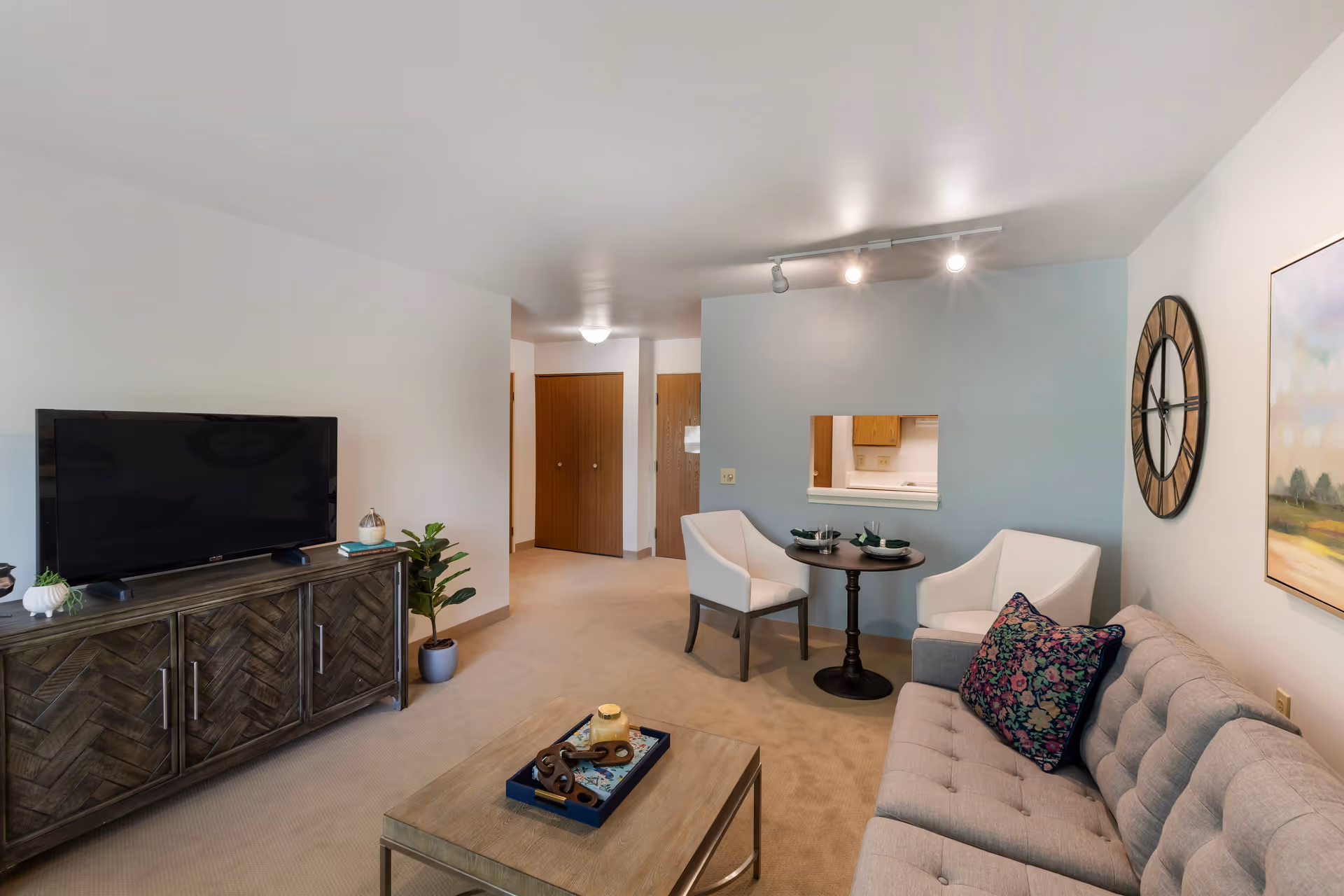 A cozy living room in a senior living facility featuring a gray tufted sofa with a floral pillow, a wooden coffee table with decorative items, a wooden TV stand with a flat-screen television, two white chairs around a small round table set for two, a large wall clock, and a painting on the wall. The room has beige carpet and light-colored walls with a small pass-through window to the kitchen.