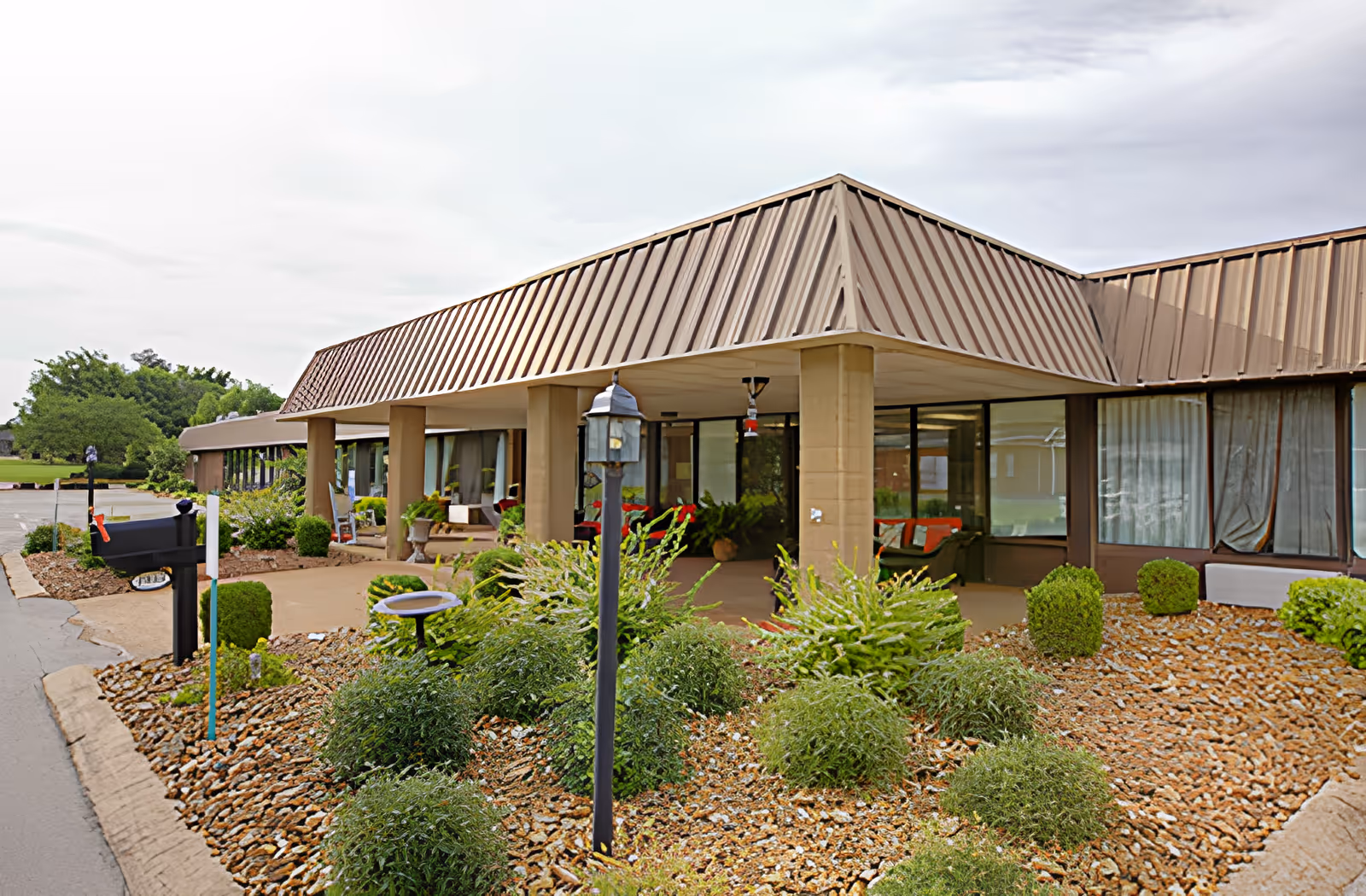 Exterior view of Ahc West Tennessee Transitional Care facility showing a single-story building with a brown metal roof and large windows. The entrance area is covered and surrounded by landscaped bushes, plants, and a rock garden. A black mailbox and a lamp post are visible near the driveway.