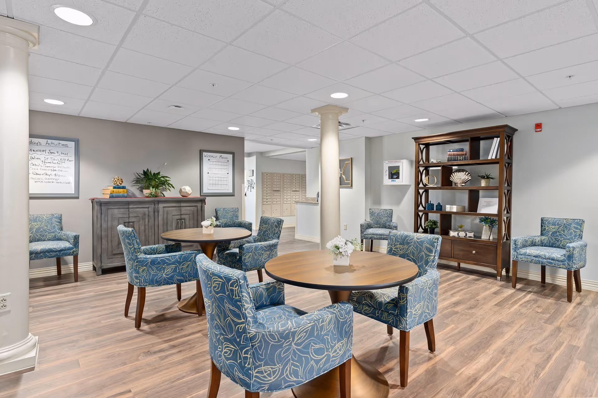 A bright and clean common area in a senior living facility featuring round wooden tables surrounded by blue patterned upholstered chairs. The room has light wood flooring, white walls, and a white ceiling with recessed lighting. There are decorative elements including a wooden bookshelf with plants and ornaments, a gray cabinet with books and plants, and framed activity schedules on the walls. Two white columns are also visible in the space.