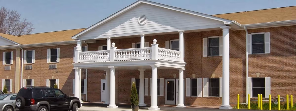 Exterior view of a two-story brick assisted living facility with white columns and a balcony at the entrance. Several windows with white shutters are visible, along with parked cars in front of the building.