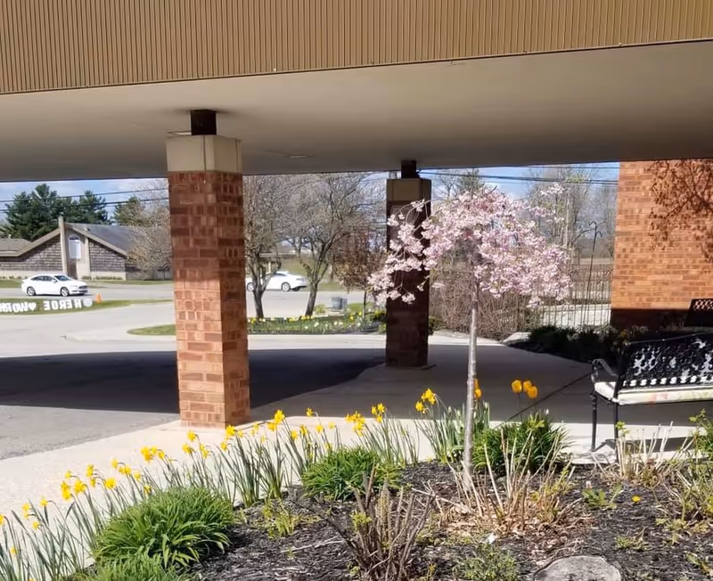 Covered driveway entrance with brick columns, a bench, a small flowering tree and daffodils in a landscaped bed.