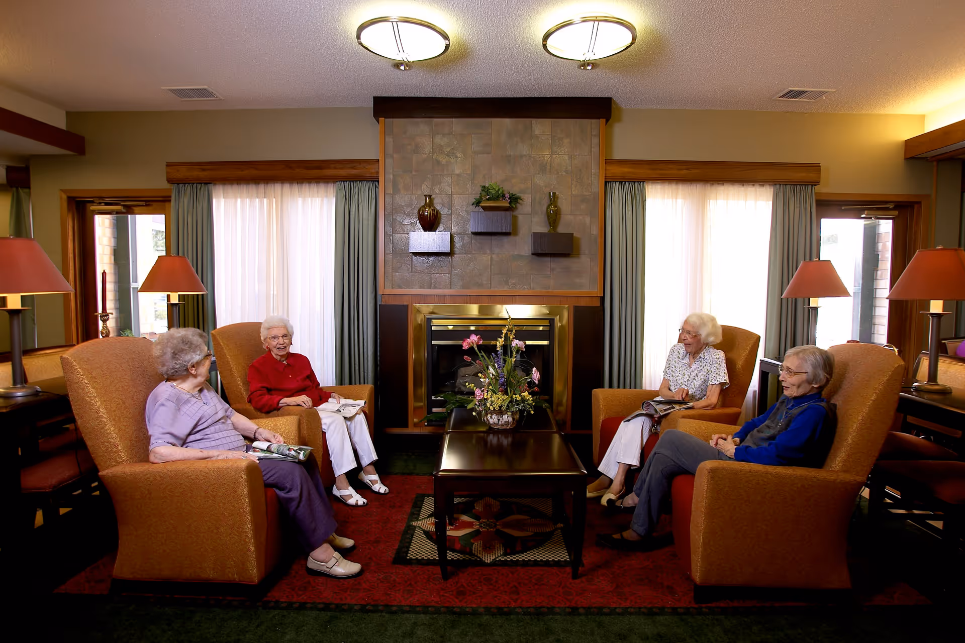 Four elderly women sitting in comfortable armchairs arranged around a wooden coffee table with a floral centerpiece in a cozy living room with a fireplace, large windows with curtains, and warm lighting from table lamps.