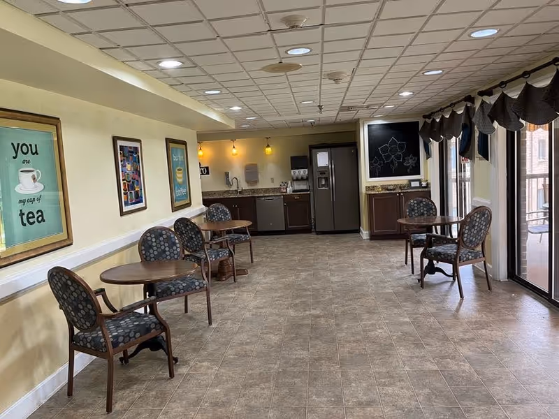 Small communal dining and coffee area with round tables and chairs, a kitchenette with refrigerator at the back, and sliding glass doors to an outdoor patio.