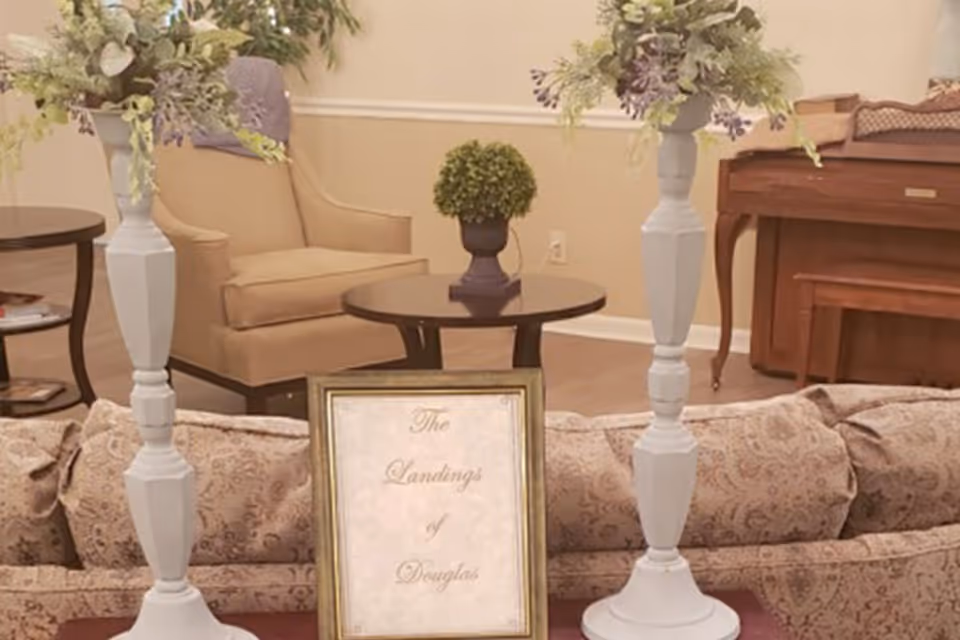 A cozy living room area with a patterned beige sofa, two tall white candle holders with floral arrangements, a small round table with a green potted plant, a beige armchair, and a wooden piano in the background. A framed sign on the sofa reads 'The Landings of Douglas'.