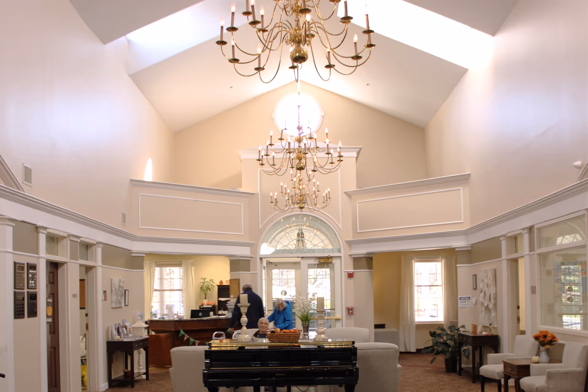 A spacious and well-lit assisted living facility common area with high vaulted ceilings and multiple elegant chandeliers. The room features a black piano in the center, surrounded by comfortable seating. There are large windows allowing natural light to fill the space, and a reception desk is visible in the background with two people near it. The walls are painted in neutral tones with white trim and decorative molding.