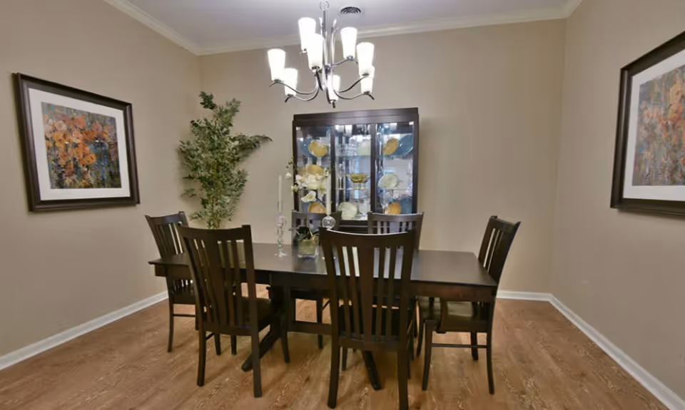 A dining room with a dark wooden table and six matching chairs. A chandelier with six lights hangs above the table. On the table are decorative items including a vase with flowers and candlesticks. Behind the table is a glass cabinet displaying plates and glassware. The room has beige walls with two framed floral paintings and a potted plant in the corner. The floor is wooden.