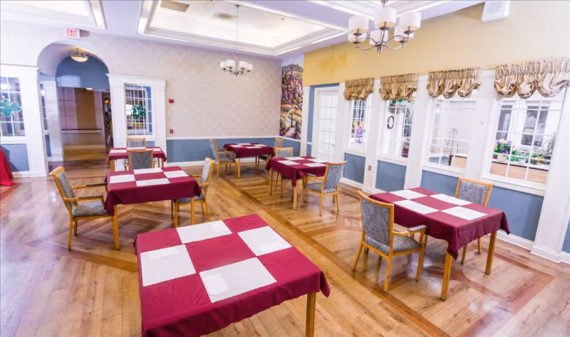A bright dining room with wooden floors and several tables covered with red tablecloths and white placemats. Each table is surrounded by wooden chairs with cushioned seats. Large windows with beige curtains allow natural light to fill the room. The walls are painted in soft colors with decorative molding and a chandelier hanging from the ceiling.