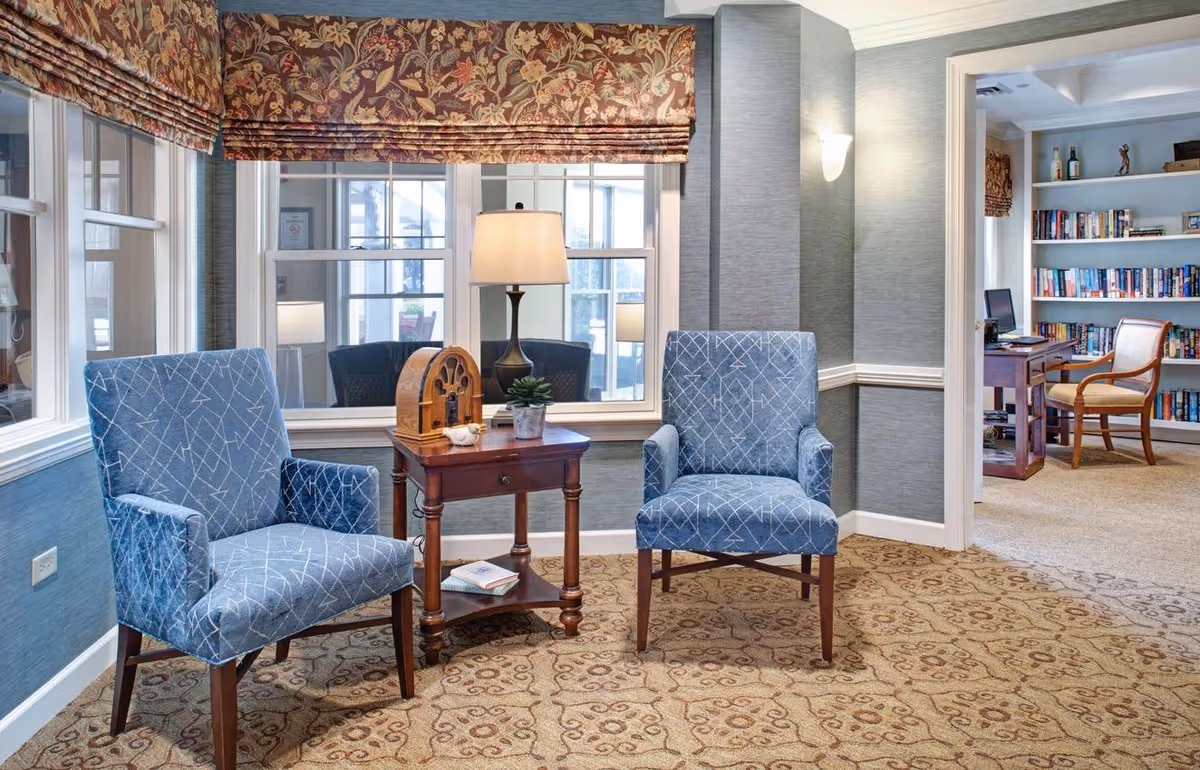 A cozy sitting area with two blue patterned armchairs and a wooden side table with a lamp, a small plant, and a decorative item. The room has large windows with floral Roman shades and a patterned carpet. In the background, there is an adjacent room with bookshelves filled with books, a wooden desk, and a chair.
