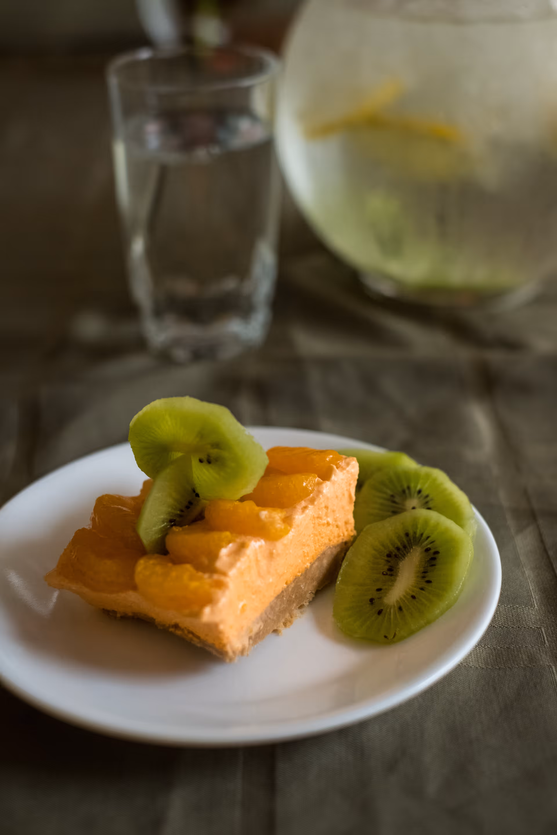 A slice of orange-colored dessert topped with mandarin orange segments and kiwi slices on a white plate, with a glass of water and a pitcher with lemon slices in the background.