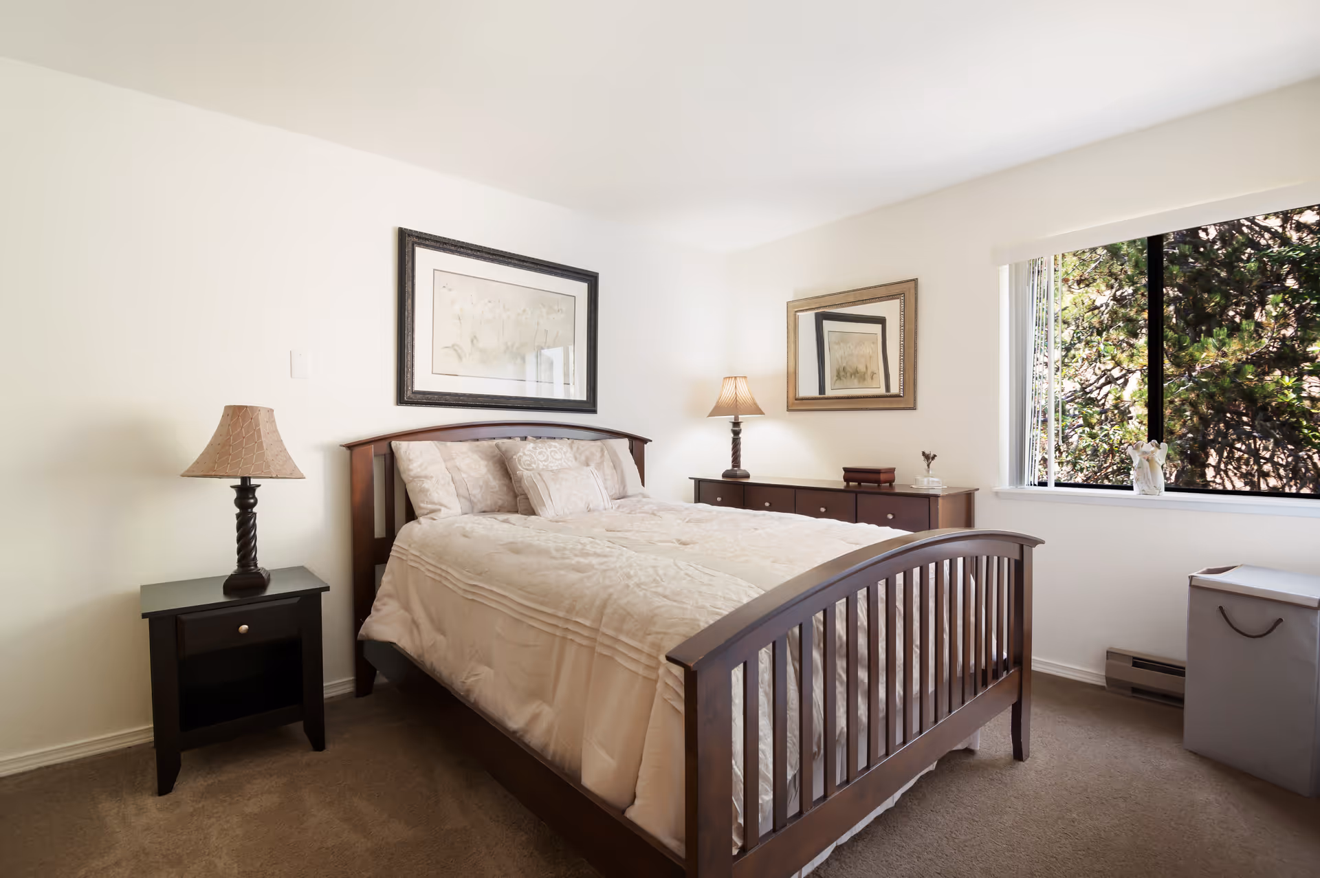 A bright bedroom with a wooden bed, nightstands and lamps, a dresser with framed artwork, and a window showing trees outside.