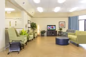 A spacious nursing home common area with light green armchairs and sofas arranged around a dark blue round ottoman. A flat-screen TV is mounted on a wooden cabinet against the far wall, with framed artwork on either side. The room has light-colored walls, wood flooring, and large windows with blue curtains allowing natural light to enter.