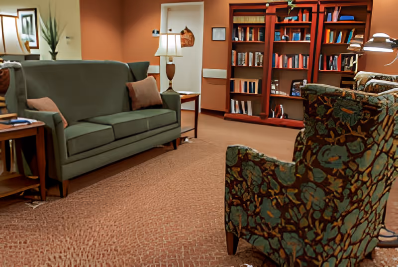 A cozy living room area with a green sofa, two patterned armchairs, a wooden bookshelf filled with books, a side table with a lamp, and warm lighting.