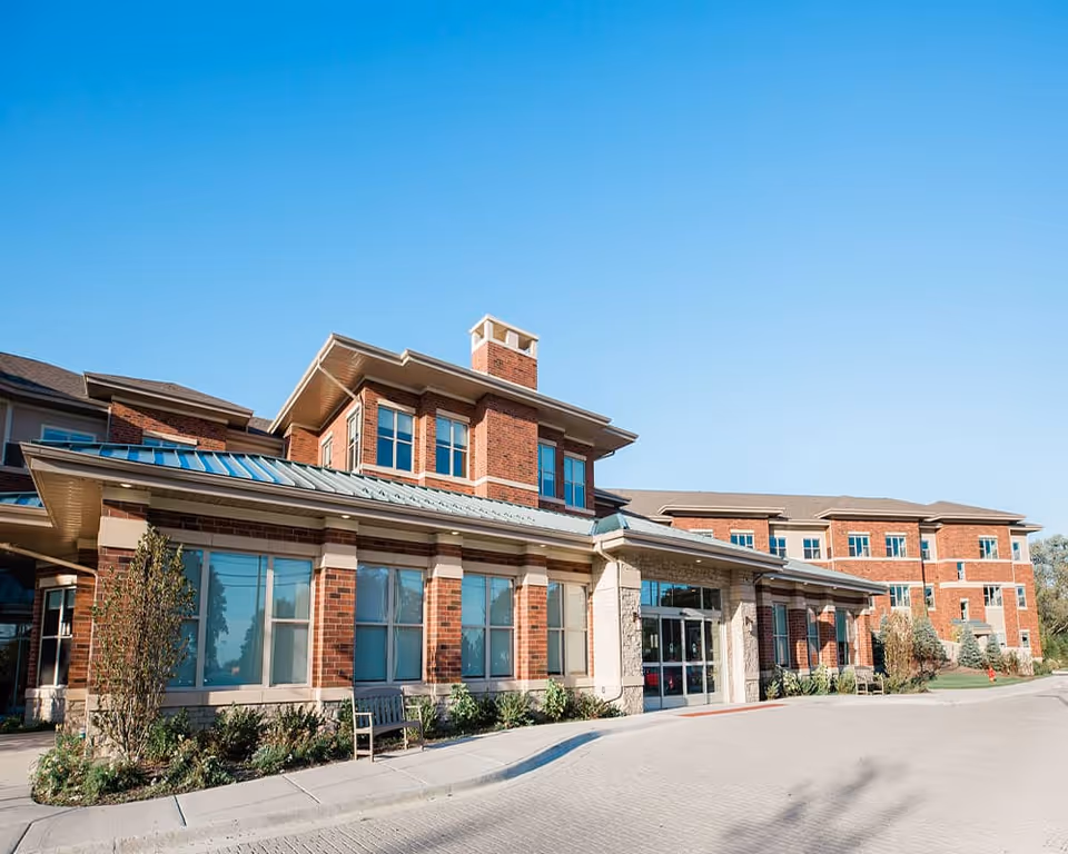 Exterior view of a senior living facility building with red brick walls, large windows, and a metal roof under a clear blue sky. There is a paved driveway and some landscaping with bushes and small trees near the entrance.