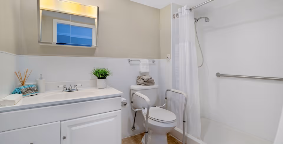 A clean and accessible bathroom with a white vanity and sink, a mirror with lighting above it, a toilet with safety rails, a stack of folded towels on top, and a walk-in shower with a white curtain and grab bar.