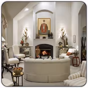 A cozy living room with a curved white sofa facing a fireplace. The fireplace is framed by a white mantel and has a fire burning inside. Above the mantel is a large portrait painting. The room features white walls, two wall sconces with candles on either side of the fireplace, and several chairs and small tables with decorative items and plants.