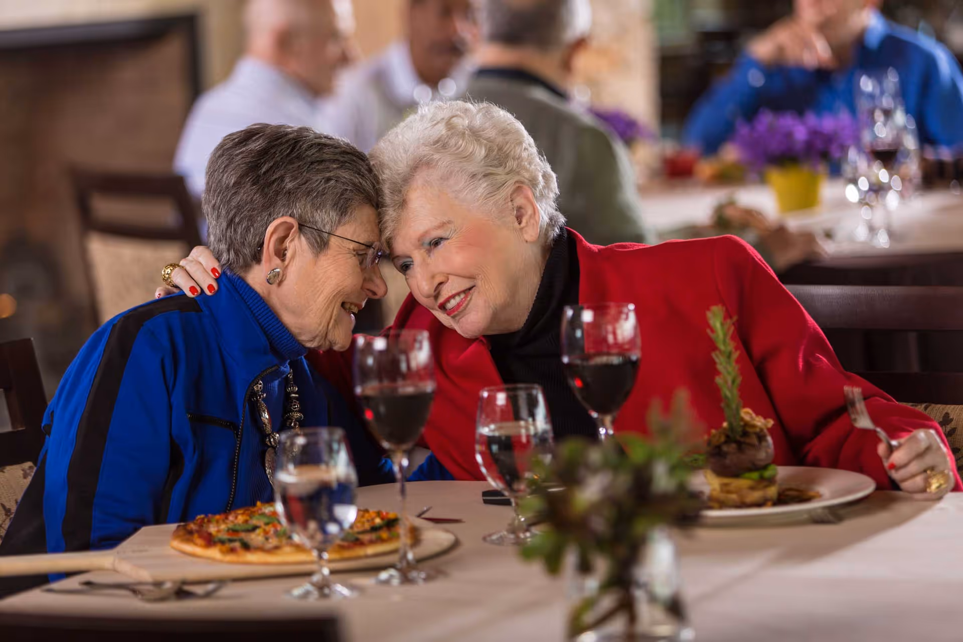 Two older women leaning together and smiling while seated at a dining table with plates and wine glasses.