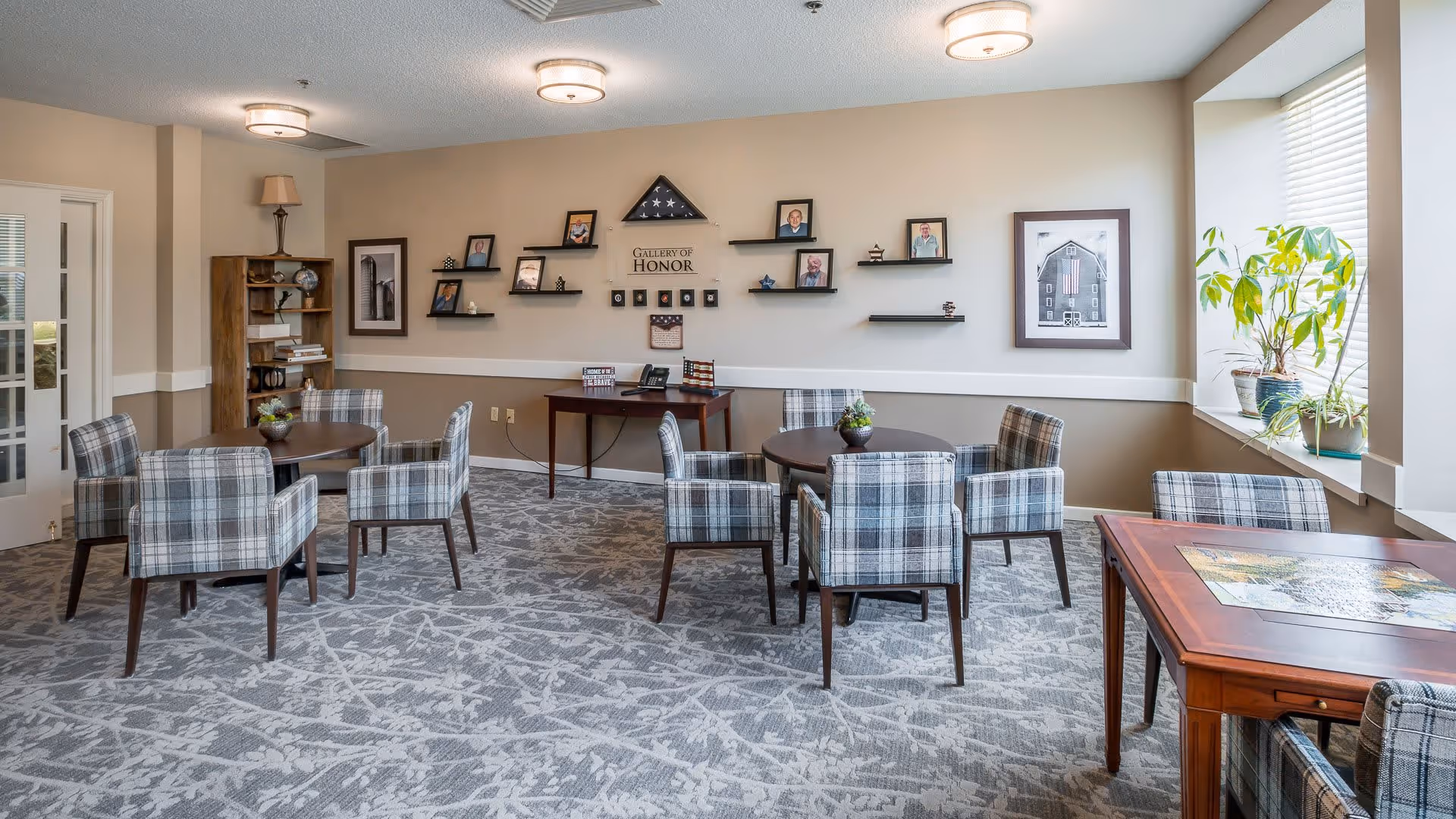 A well-lit common area with multiple round tables surrounded by plaid upholstered chairs. The walls are decorated with framed photos and a 'Gallery of Honor' display. There is a window with plants on the windowsill allowing natural light into the room. The carpet has a modern pattern, and the ceiling has round light fixtures.