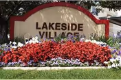 A large sign reading 'LAKESIDE VILLAGE' surrounded by a colorful flower bed with red, white, and purple flowers, set on a grassy area with trees and a building partially visible in the background.