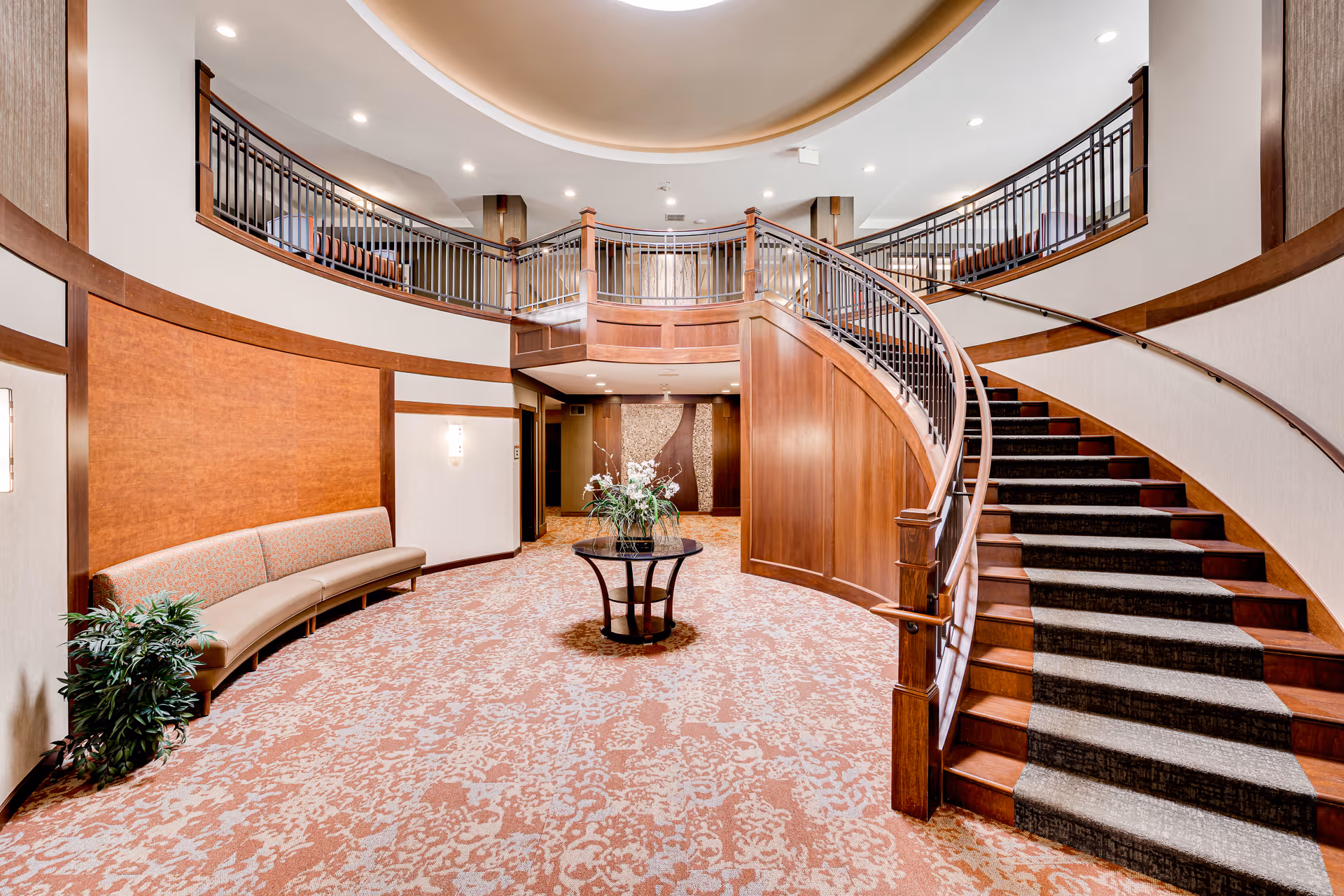 A spacious interior area featuring a curved wooden staircase with carpeted steps leading to an upper level with a railing. The room has patterned carpet flooring, a curved bench along the left wall, a round table with a floral arrangement in the center, and soft lighting from wall sconces and ceiling lights.