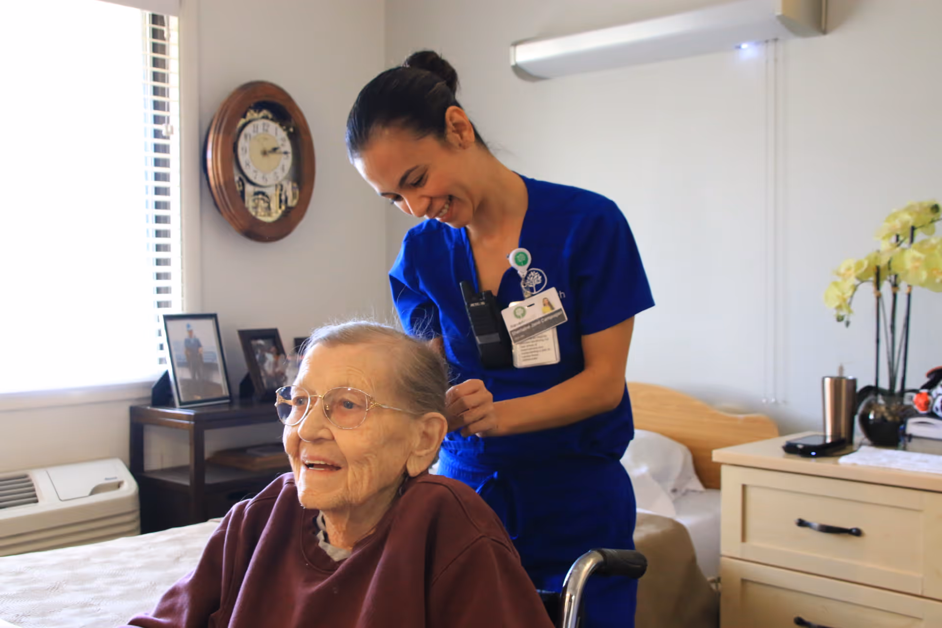 A smiling healthcare worker in blue scrubs is standing behind an elderly woman in a wheelchair inside a bedroom. The elderly woman is wearing glasses and a maroon sweater. The room has a bed, a nightstand with a plant and personal items, a wall clock, and framed photos on a small table near a window with blinds.