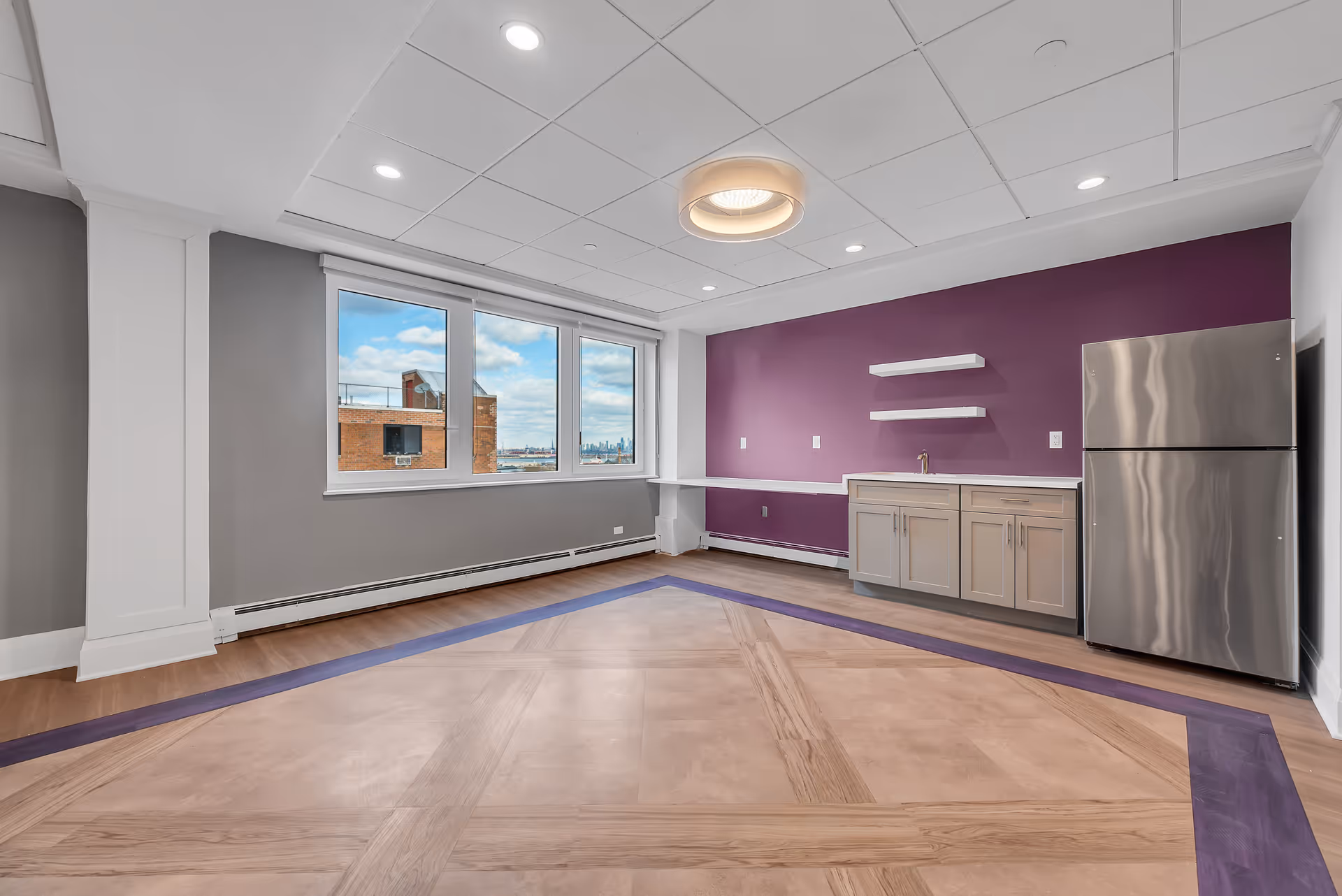 A modern interior room with a large window showing a cityscape outside. The room features a purple accent wall with two white floating shelves, a small sink with cabinetry, and a stainless steel refrigerator. The floor has a decorative wooden pattern with a purple border. The ceiling has recessed lighting and a circular light fixture.