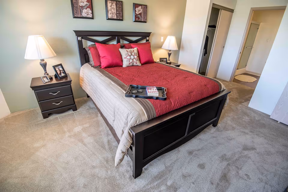 A neatly made bed with a dark wooden frame and red and beige bedding in a bedroom. There are two bedside tables with lamps and framed photos on each side of the bed. Three floral paintings hang on the wall above the headboard. The room has beige carpet flooring and an open doorway leading to a closet and bathroom area.