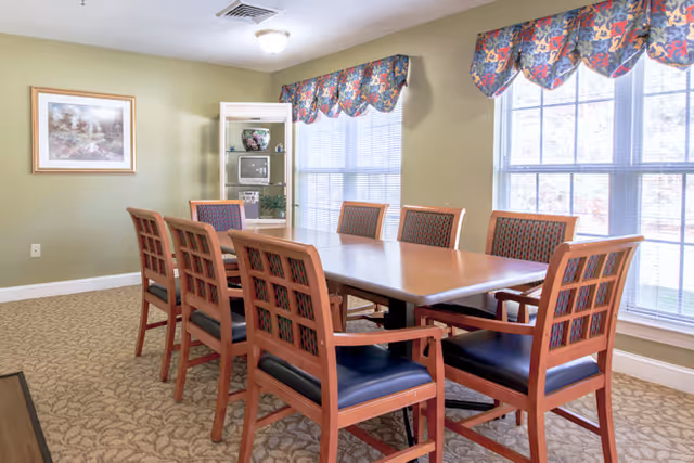 A dining room with a rectangular wooden table surrounded by eight wooden chairs with blue cushioned seats. The room has light green walls, two large windows with floral valances, a framed picture on the wall, and a glass display cabinet with decorative items.