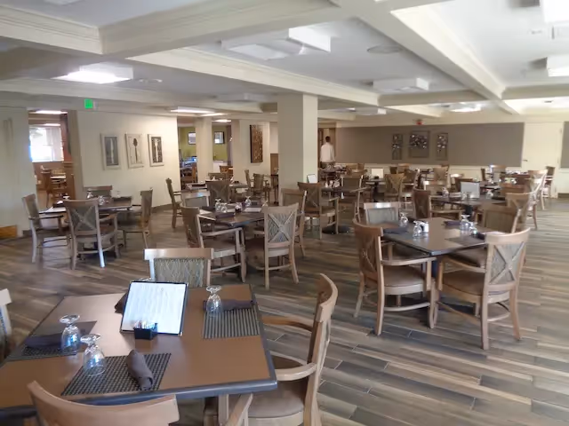A spacious dining room in an assisted living center with multiple wooden tables and chairs arranged neatly. Each table is set with placemats, napkins, and upside-down glasses. The room has a wooden floor and neutral-colored walls with framed artwork. A person is visible in the background near the far wall.