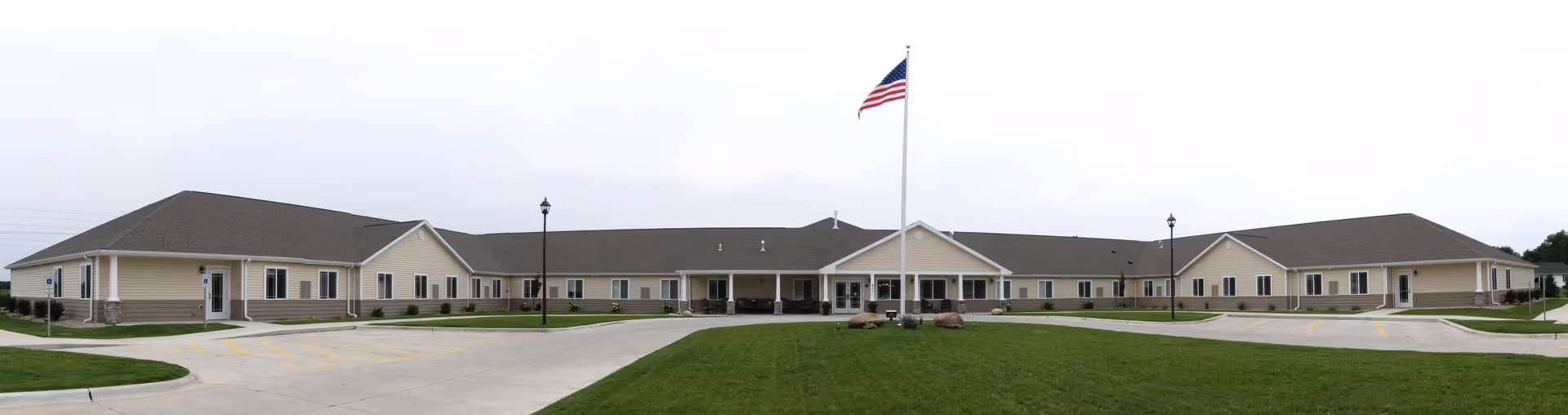 Wide panoramic view of a single-story senior living facility building with beige siding and a dark roof. The building is U-shaped with a central entrance under a covered porch. An American flag flies on a flagpole in front of the entrance, and there are street lamps and paved driveways surrounding the building. The sky is overcast.