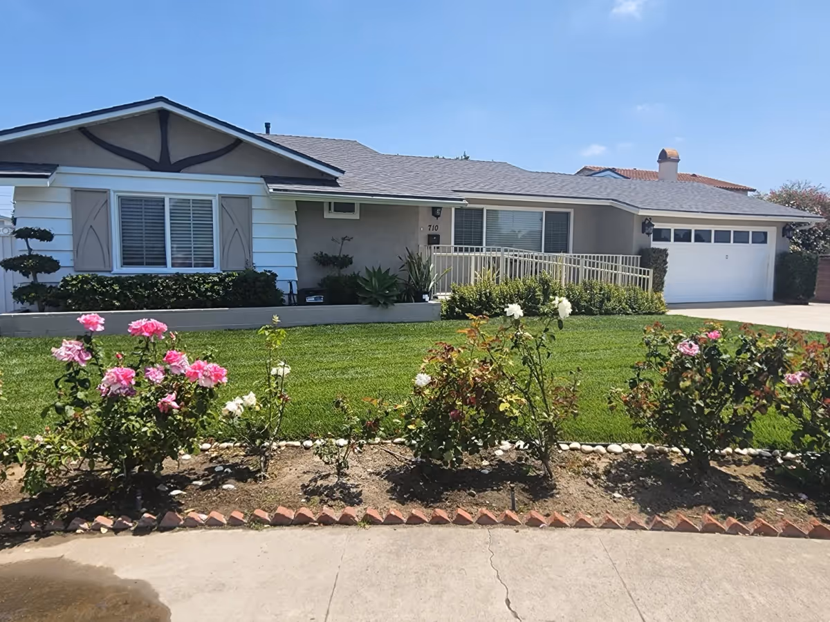 Front exterior view of a single-story residential building with a well-maintained lawn and flower bed featuring pink and white roses. The house has a gray roof, beige walls, white window shutters, and a white garage door. There is a ramp leading to the front door.