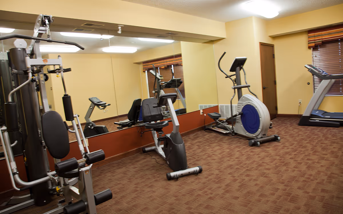 A small fitness room with exercise equipment including a multi-station gym machine, a recumbent exercise bike, an elliptical machine, and a treadmill. The room has beige walls, a large mirror on one wall, brown carpeted floor, and a window with brown blinds and a striped valance.
