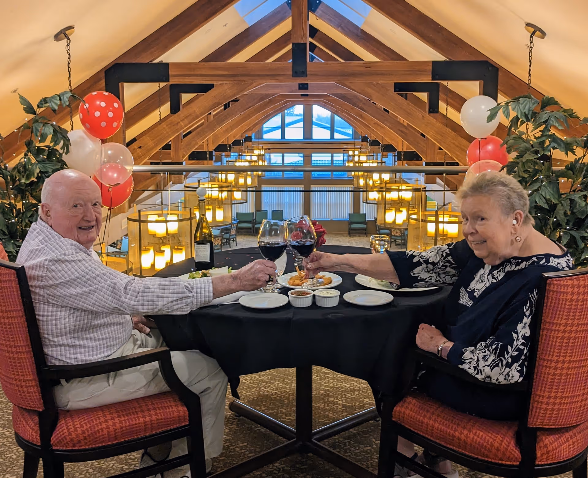 An elderly man and woman sitting at a round table with a black tablecloth, toasting with glasses of red wine in a warmly lit dining area with wooden beams and hanging lights. There are red and white balloons and green plants around them.