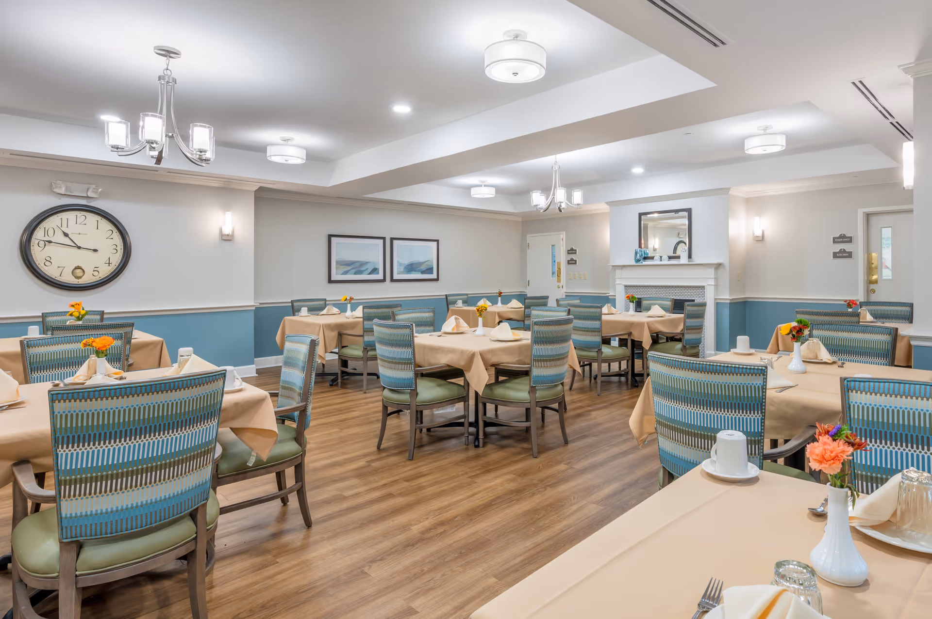 Bright dining room with tables set with beige tablecloths, patterned chairs, and small floral centerpieces.