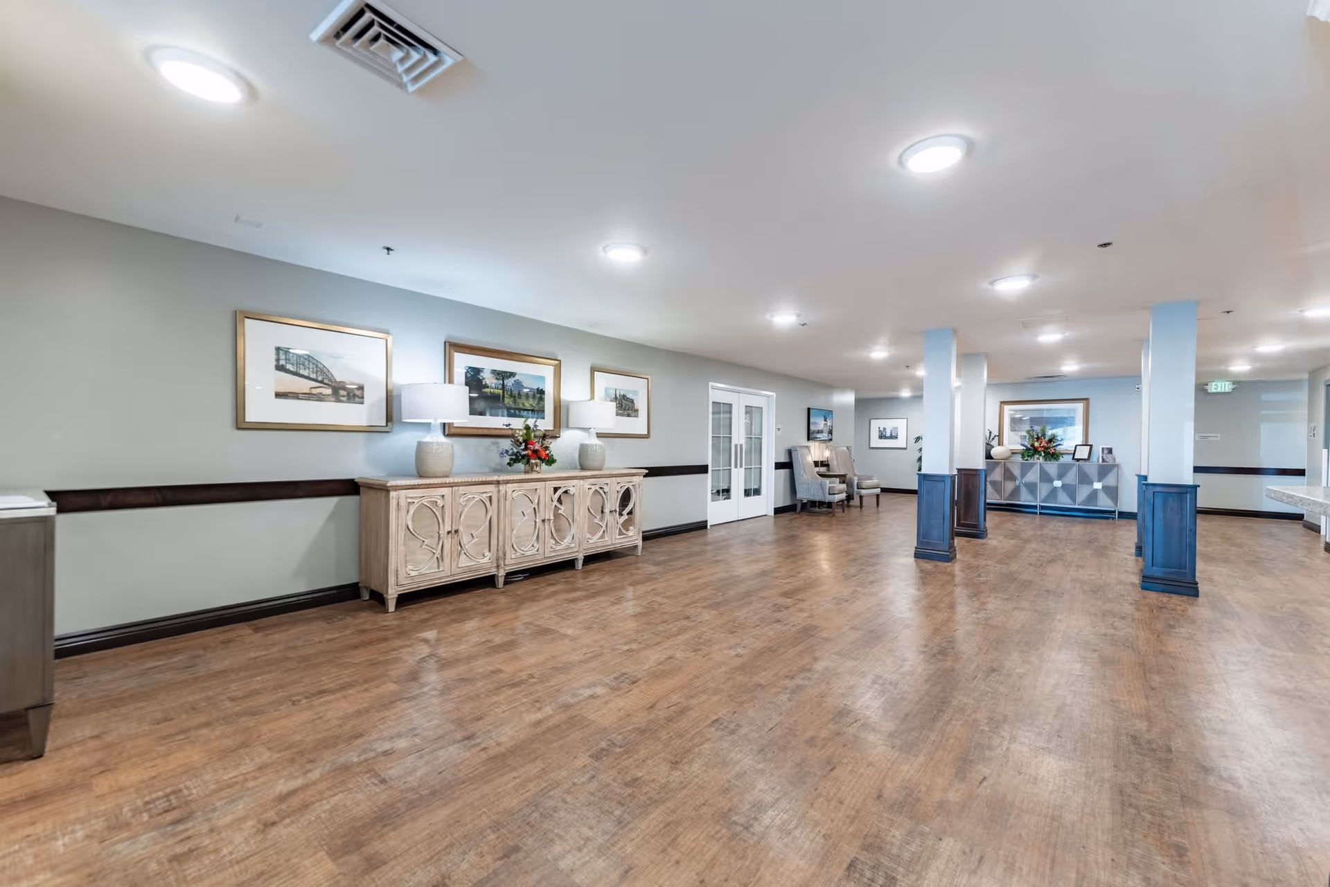 A spacious, well-lit interior hallway or common area with wooden flooring, light gray walls, and several framed pictures on the walls. There are decorative cabinets with lamps and flower arrangements, a couple of armchairs near a set of glass double doors, and several columns painted white with dark wood bases.