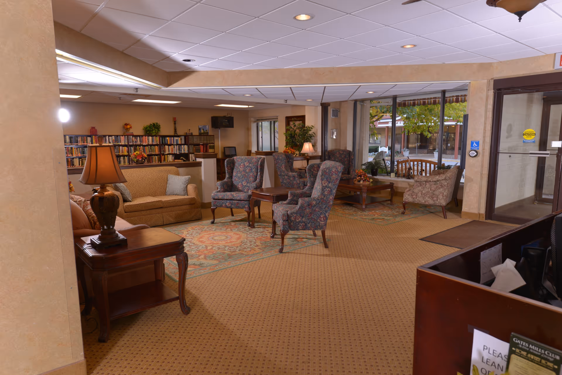 A senior living facility lobby with sofas, patterned armchairs, tables, a bookcase and the glass entrance visible.