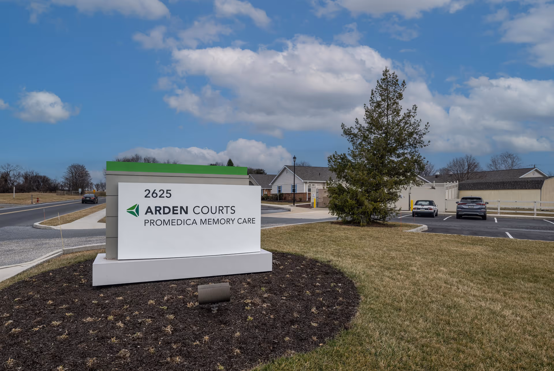 Outdoor view of Arden Courts ProMedica Memory Care Community sign with address 2625, situated on a landscaped area with grass and mulch. In the background, there is a parking lot with a few cars, a tree, and residential-style buildings under a partly cloudy sky.