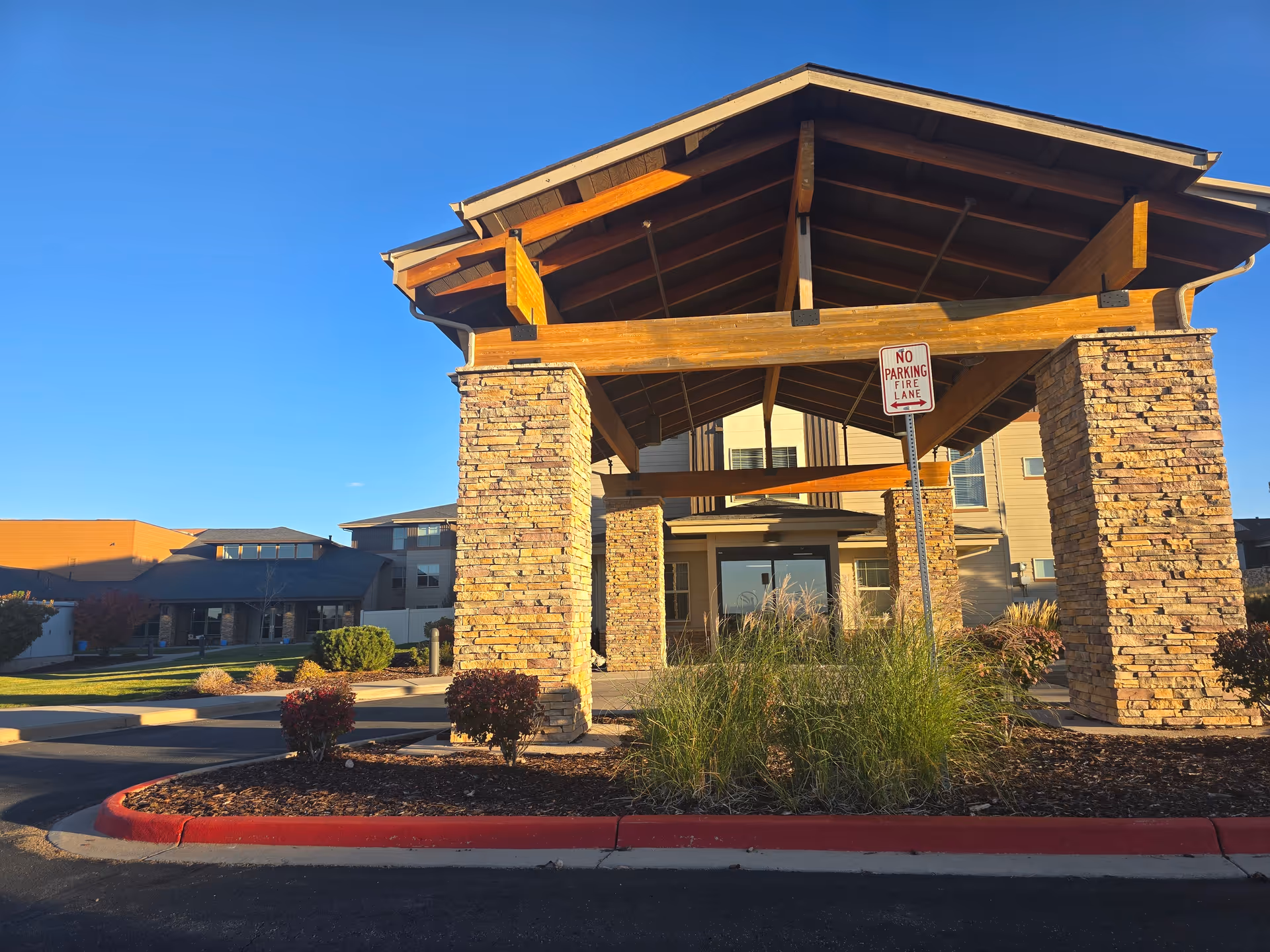 Entrance of a senior living facility with a covered drop-off area supported by stone pillars and wooden beams, landscaped with bushes and grass under a clear blue sky.
