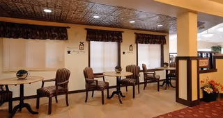 Interior view of a common area in a senior living facility with several round tables and striped upholstered chairs arranged along a wall with three windows covered by valances and blinds. The ceiling has a decorative pattern, and there are wall decorations and a column dividing the space. A small flower arrangement is visible on one table.