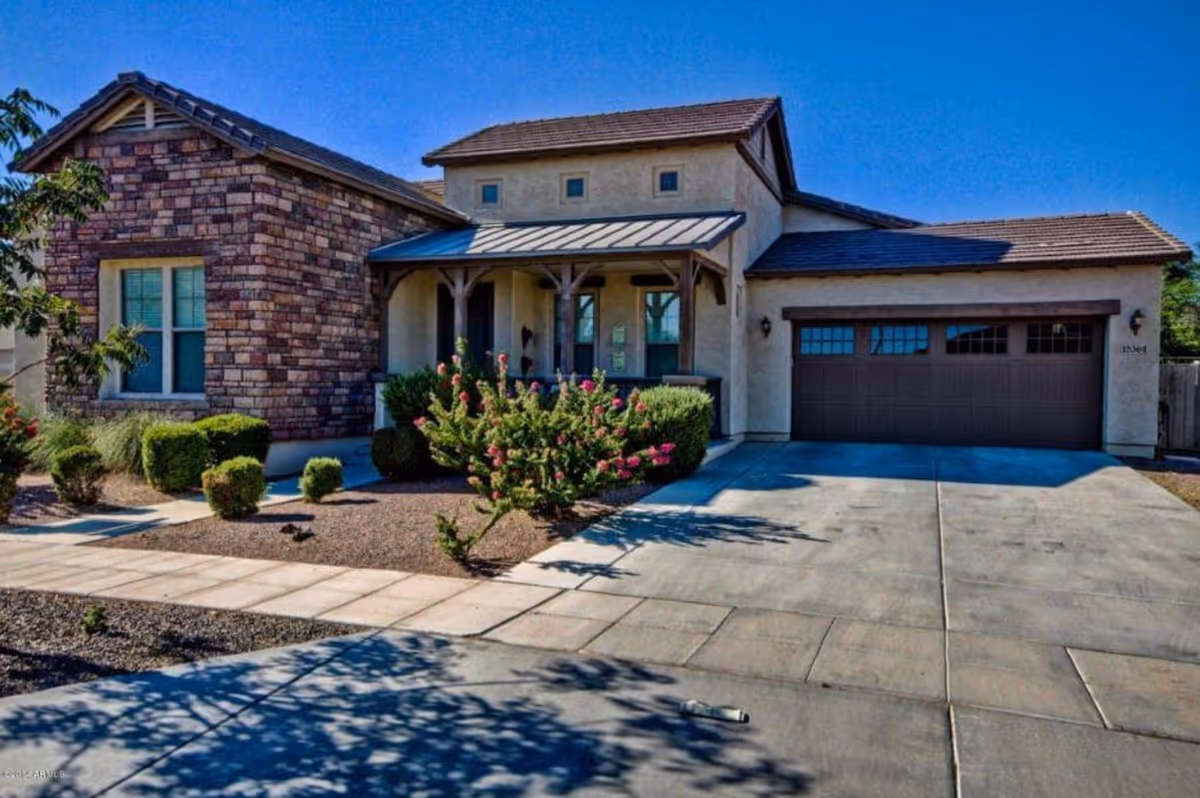 Front exterior view of a single-story house with a combination of stone and stucco walls, a covered porch with wooden beams, a two-car garage with brown doors, and a concrete driveway. The landscaping includes small bushes and flowering plants under a clear blue sky.