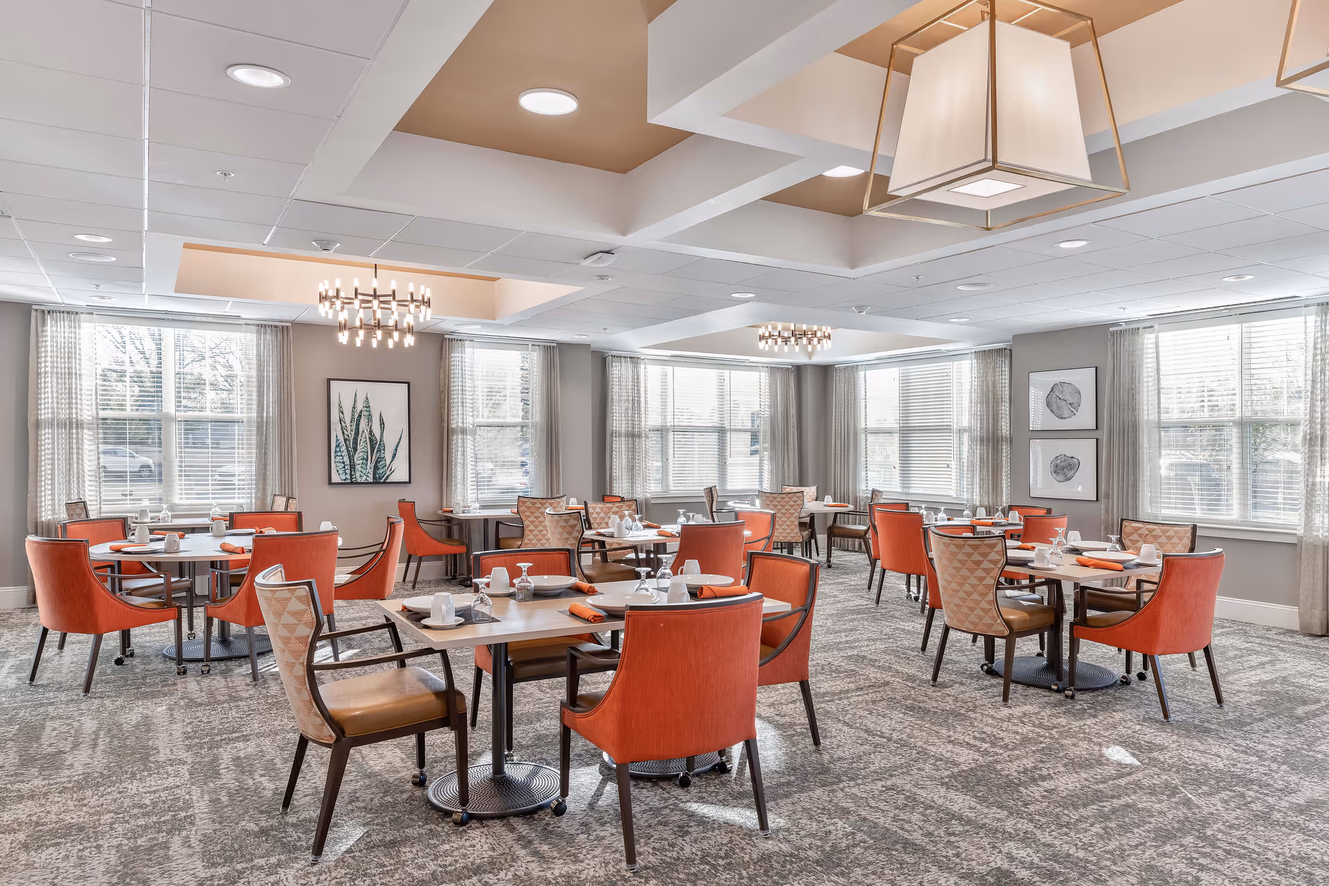 A bright and spacious dining room with multiple tables set for meals. The room features large windows with sheer curtains allowing natural light to fill the space. The tables are surrounded by orange and patterned cushioned chairs. Modern light fixtures hang from the ceiling, and framed botanical artwork decorates the walls.