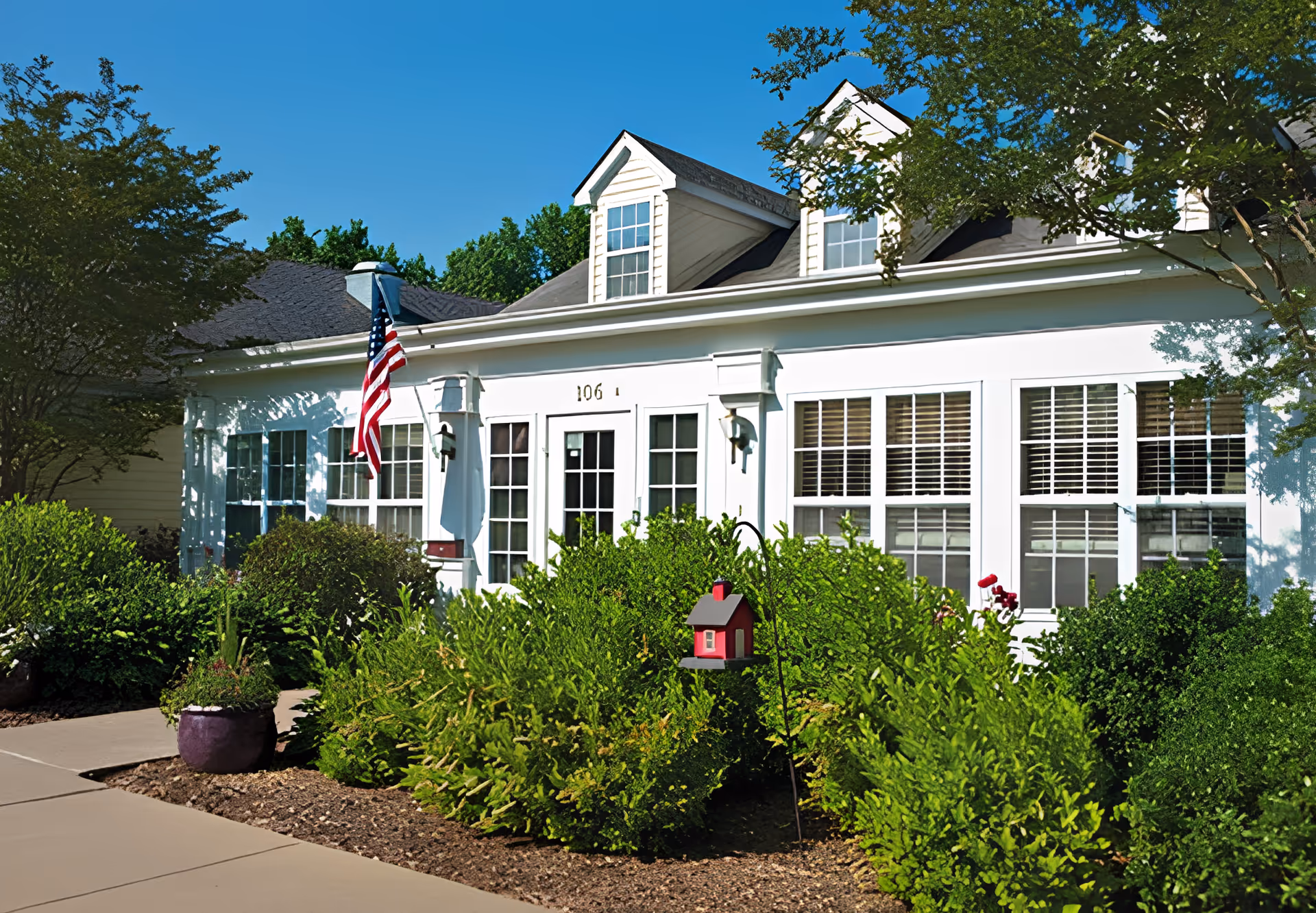 Exterior view of a white building with multiple windows and dormer windows on the roof. There is an American flag mounted near the entrance, surrounded by green bushes and trees under a clear blue sky.
