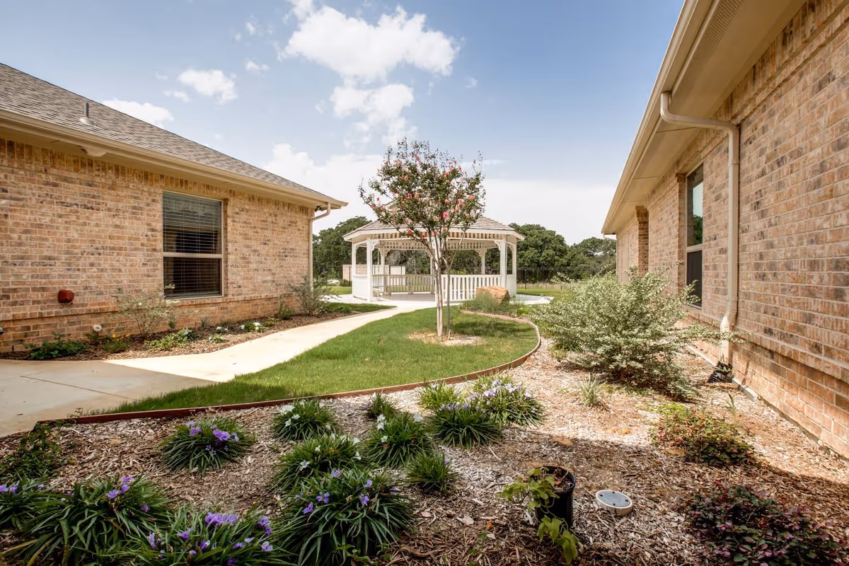 Outdoor garden area between two brick buildings with a concrete walkway, green grass, various plants, and a white gazebo in the background under a partly cloudy sky.