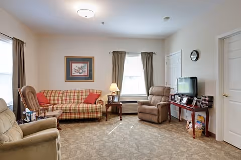 A cozy living room with a plaid sofa, two armchairs, a side table with a lamp, a window with curtains, a wall clock, and a TV on a wooden console table. The room has beige walls and carpeted floor.