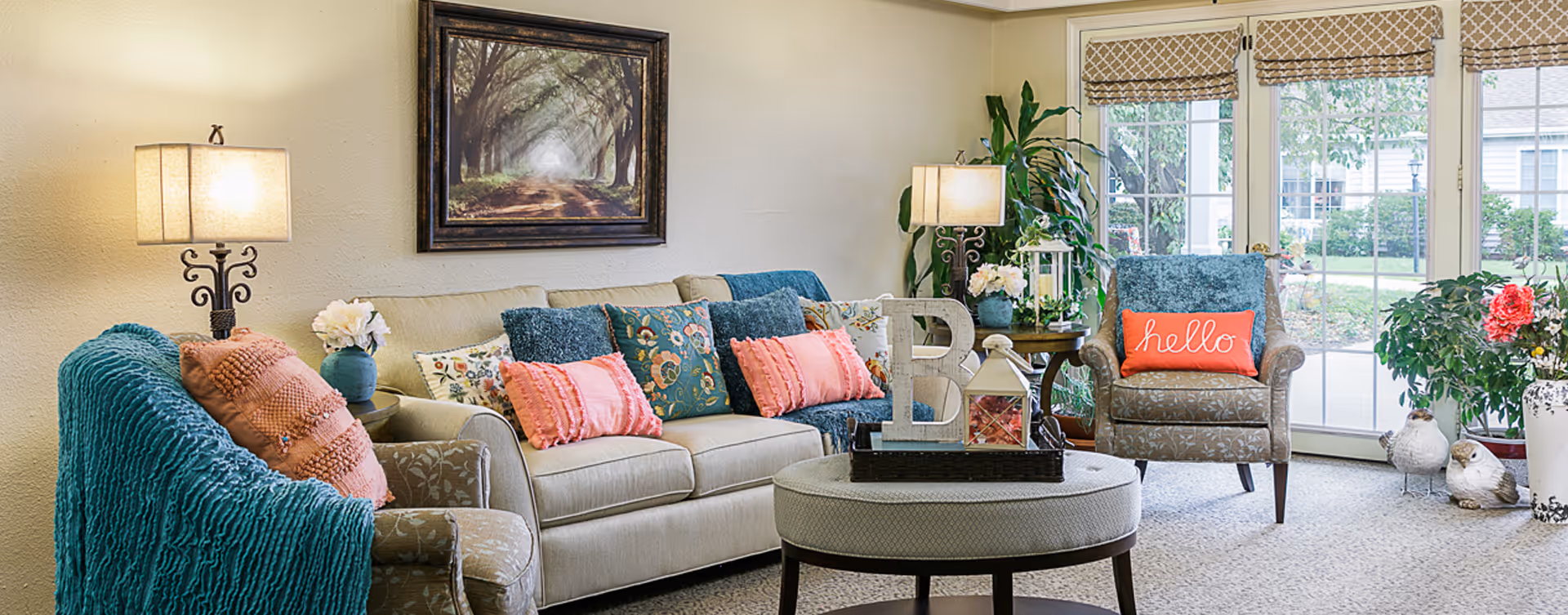 Bright communal living room with a sofa and armchairs dressed in colorful pillows and throws, a round ottoman coffee table, lamps, potted plants, and large windows.
