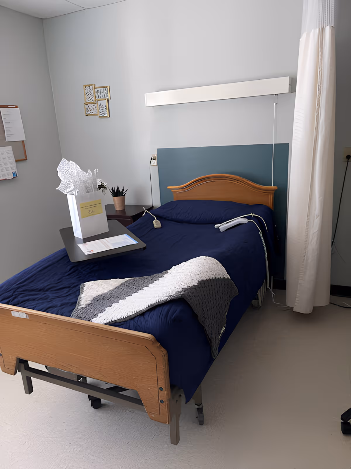 A single hospital-style bed with a wooden headboard and footboard, covered with a dark blue blanket and a gray and white knitted throw. A small overbed table holds a white gift bag with tissue paper and some papers. The room has light gray walls, a small potted plant on a side table, a wall-mounted light fixture above the bed, and a white privacy curtain partially drawn to the side.