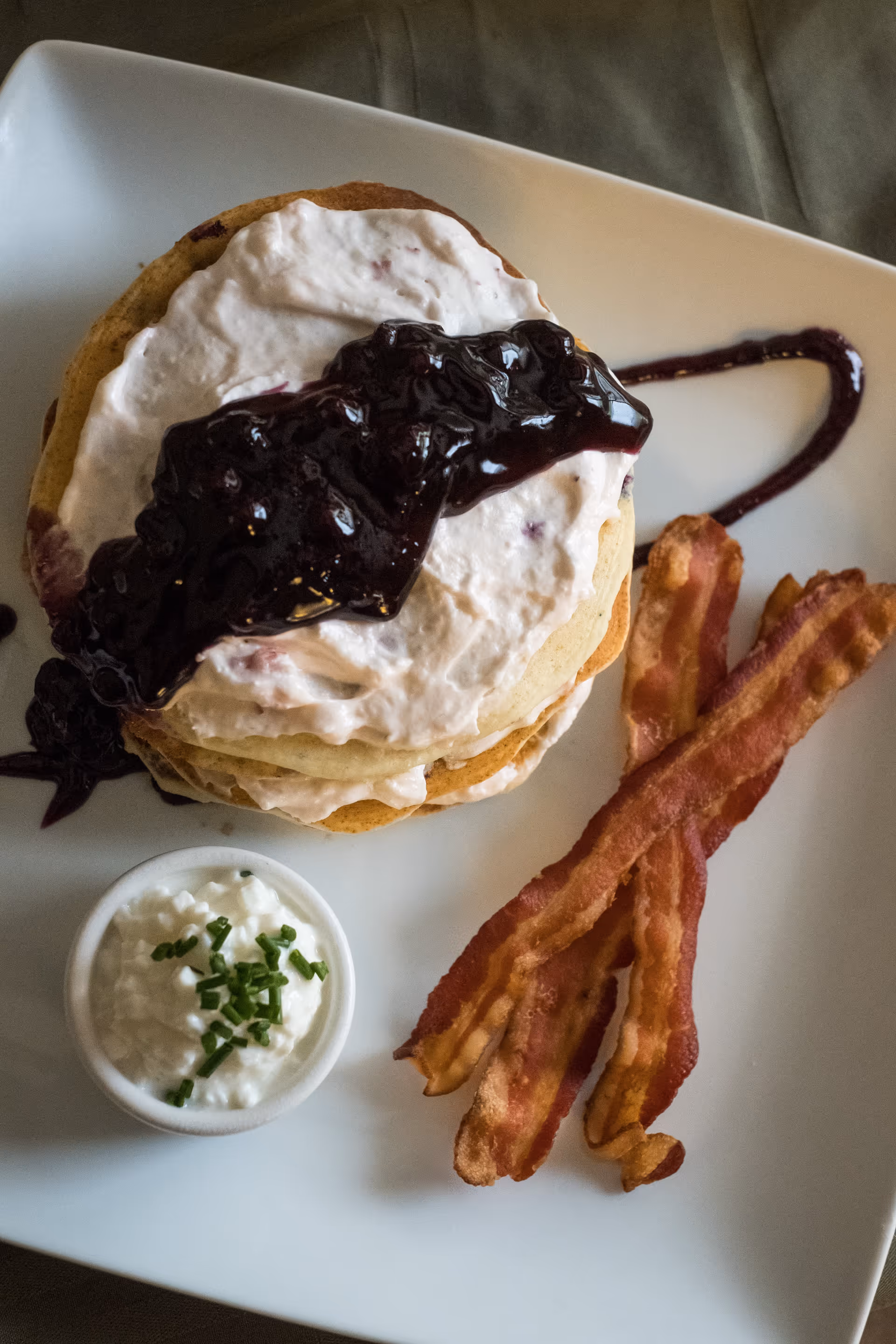 A plate with a stack of pancakes topped with a creamy spread and blueberry sauce, served with two strips of crispy bacon and a small cup of cottage cheese garnished with chopped chives.
