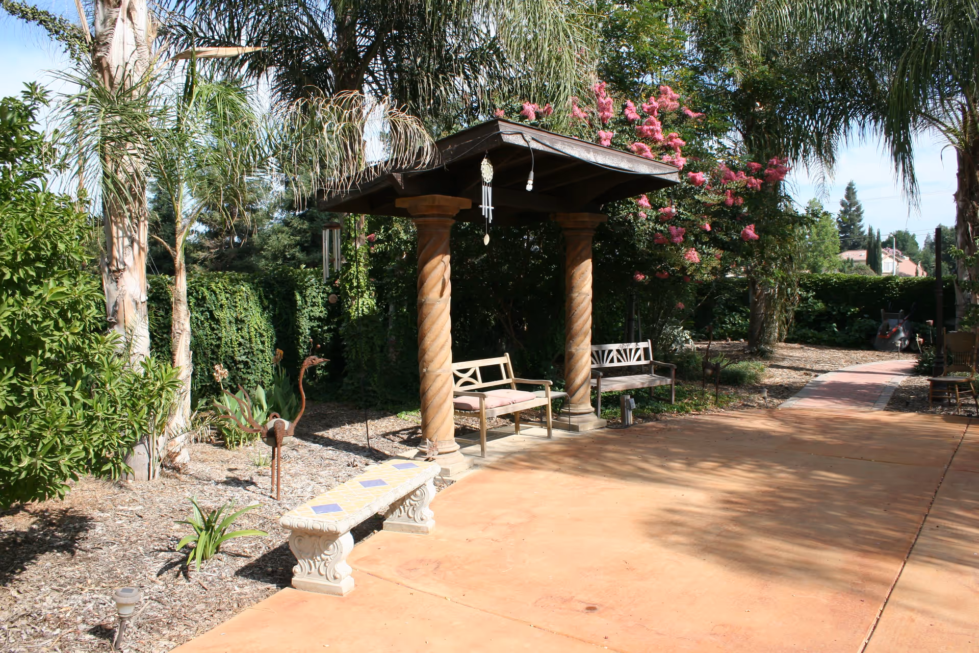 Outdoor garden area with a paved walkway, two wooden benches under a small wooden pergola with twisted columns, a decorative stone bench with a tiled top, surrounded by various trees and plants including palm trees and flowering bushes.