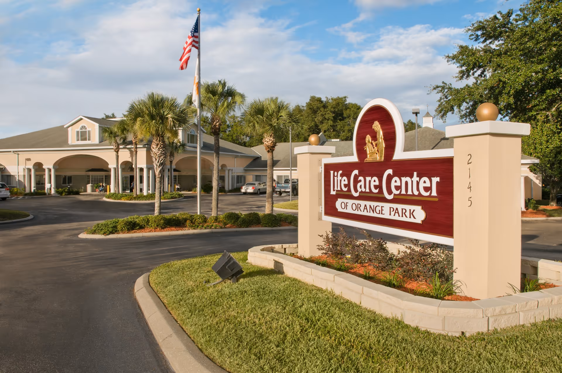 Front entrance of the Life Care Center of Orange Park with a large sign, driveway, palm trees and flagpoles.